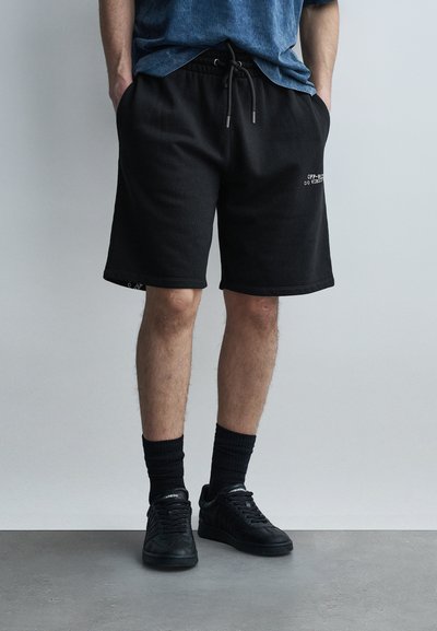 Homme portant un short noir avec cordon de serrage, des chaussettes noires montantes et des baskets noires, debout devant un fond uni gris clair.