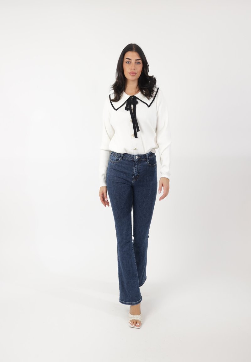 Suéter blanco con un gran collar negro y un lazo, combinado con jeans acampanados azul oscuro y sandalias claras, sobre un fondo blanco.