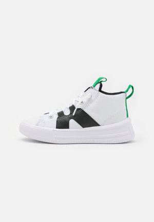 Zapatillas altas con parte superior de lona blanca, detalles en negro y un lazo verde en el talón. Suela de goma texturizada para ofrecer agarre y durabilidad.