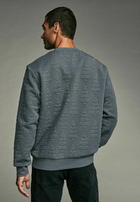 Gray sweatshirt featuring an embossed tree pattern, textured fabric, crew neck, and ribbed cuffs and hem. Worn by a person viewed from behind.