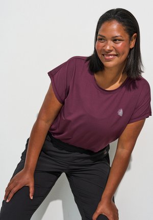 Lächelnde Frau mit glatten schwarzen Haaren, die ein violettes kurzärmeliges Shirt und schwarze Hosen trägt und sich nach vorne lehnt mit den Händen auf den Knien.