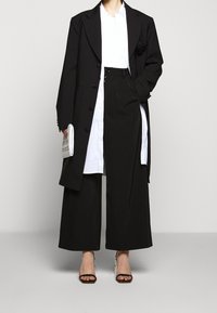 Abrigo largo negro sobre una camisa blanca, combinado con pantalones anchos negros y tacones negros con tiras. Bolso de mano blanco con texto impreso.