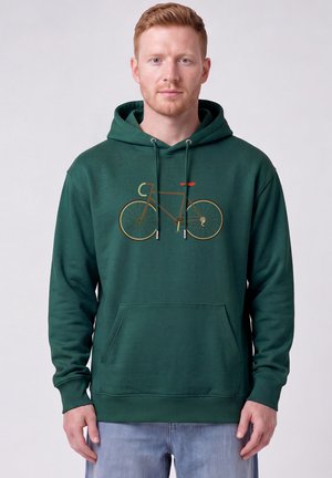 Jeune homme à la peau claire et aux cheveux roux portant un sweat à capuche vert foncé avec un motif de vélo marron et un jean bleu clair, debout devant un fond uni.