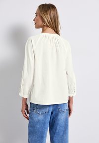 Vit, löst sittande topp med trekvartslånga ärmar, med plisseringar på baksidan och knappdetaljer på ärmsluten. Toppad med blå jeans.
