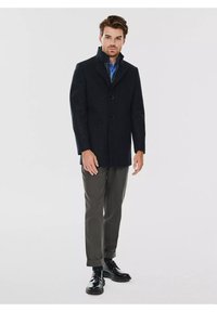 Czarny płaszcz wełniany z wysokim kołnierzem, trzema guzikami z przodu i dopasowanym krojem. Połączony z szarymi spodniami i błyszczącymi czarnymi butami.