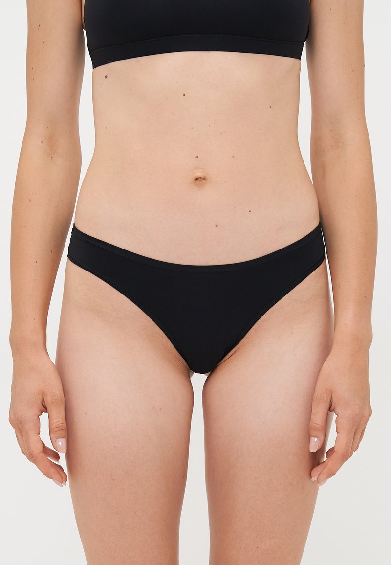 Mujer con bikini negro, mostrado desde la mitad del muslo hasta debajo del pecho, de pie contra un fondo liso.
