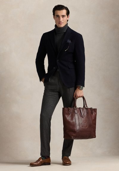 Homme en blazer sombre, col roulé gris, pantalon gris, mocassins marron, tenant un grand sac fourre-tout en cuir marron, debout devant un fond neutre.