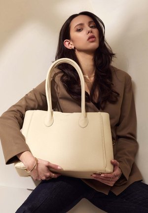 Beige leren tote bag met twee ronde handvatten, gestepte afwerking en gouden hardware accenten. Model houdt de tas tegen een neutrale achtergrond.