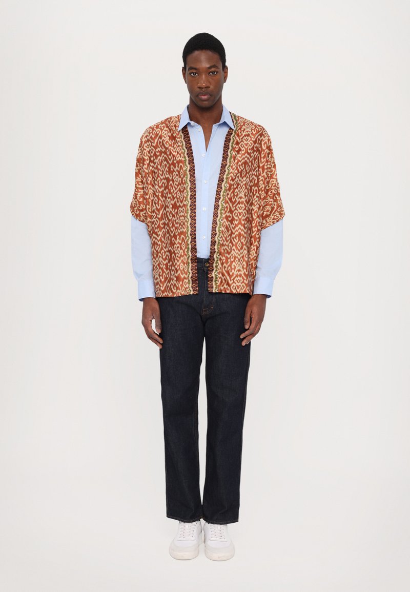 Camisa corta de manga corta a rayas en naranja y crema sobre una camisa azul claro de botones, combinada con jeans oscuros y zapatillas blancas.