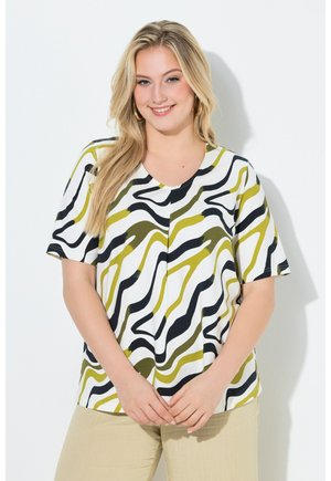 Donna sorridente con capelli lunghi e biondi che indossa orecchini a cerchio dorati, una maglietta bianca con strisce ondulate nere e verde oliva e pantaloni beige chiaro.