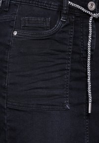 Jean en denim noir avec une finition texturée, une poche avant avec des détails de couture et un cordon de taille noir et blanc.