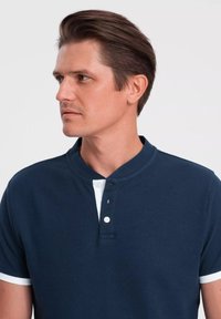 Polo shirt blu navy con colletto, caratterizzato da un'applicazione di bottoni bianchi e dettagli bianchi sui bordi delle maniche. Tessuto strutturato, maniche corte.