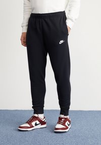 Svarta Nike-joggers med resårmidja, avsmalnande ben och vitt märke på vänster lår, kombinerat med röda och vita sportskor.