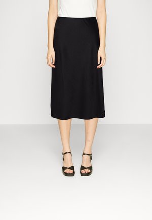 Femme portant une jupe midi noire, un haut blanc et des sandales à talons noires, debout devant un fond clair uni.