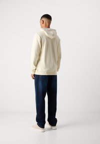 Helle Creme Kapuzenjacke mit verstellbarem Kapuzen, gerippten Bündchen, kombiniert mit dunkelblauen, lockeren Jeans und beigen Sneakern. Glatte Stoffoberfläche.