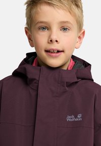 Veste imperméable bordeaux avec col haut et capuche. Elle présente une doublure intérieure rose vif et un logo blanc sur le côté gauche de la poitrine.