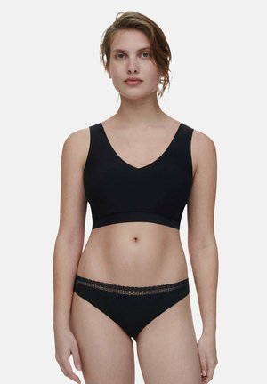 Vrouw die een zwart mouwloos crop top en zwarte bikini-ondergoed met kanten rand draagt, staand tegen een effen lichte achtergrond.