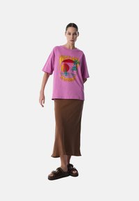 T-shirt over sized rosa con design grafico, gonna midi marrone e sandali neri scivolati con borchie. Materiale in cotone, vestibilità comoda e texture liscia.