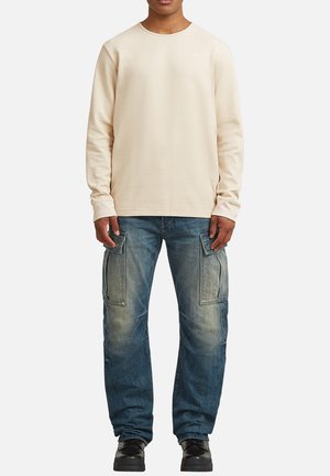 Mand iført en cremefarvet, langærmet wafflestrikket skjorte, falmede blå cargobukser med store sidelommer og sorte sko står mod hvid baggrund.