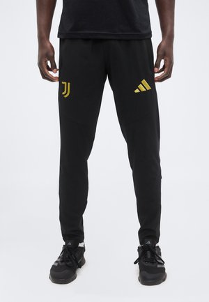 Mand iført sorte atletikbukser med guld Juventus- og Adidas-logoer, kombineret med sorte Adidas løbesko.