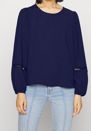Navy blue blouse with a round neckline, long sleeves, puffed shoulders, and lace detailing on the cuffs. Soft, lightweight fabric.