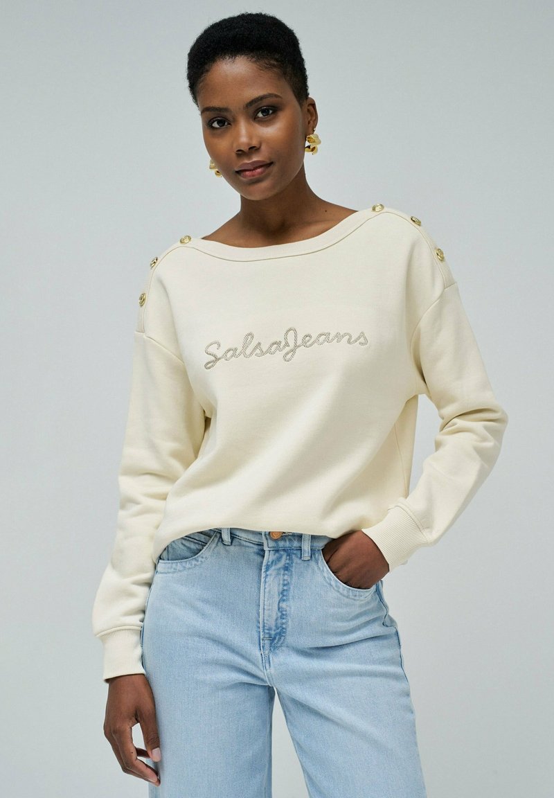 Sweatshirt crème avec "SalsaJeans" brodé, décoré de boutons dorés sur les épaules, coupe décontractée et poignets côtelés. Assorti avec un jean bleu clair.