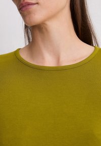 Olivgrünes, geripptes T-Shirt mit rundem Halsausschnitt, kurzen Ärmeln und einer glatten Textur. Details umfassen feine Nähte entlang des Halsausschnitts.