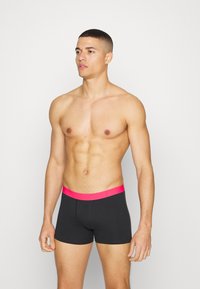 Men's black boxer briefs with a bright pink waistband. Smooth fabric, fitted design, mid-thigh length. Model displays side stance.