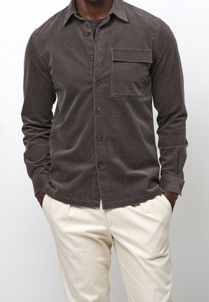 Man draagt een donkerbruine blouse met knopen en borstzak en crèmekleurige broek, handen in de zakken, staand tegen een effen achtergrond.