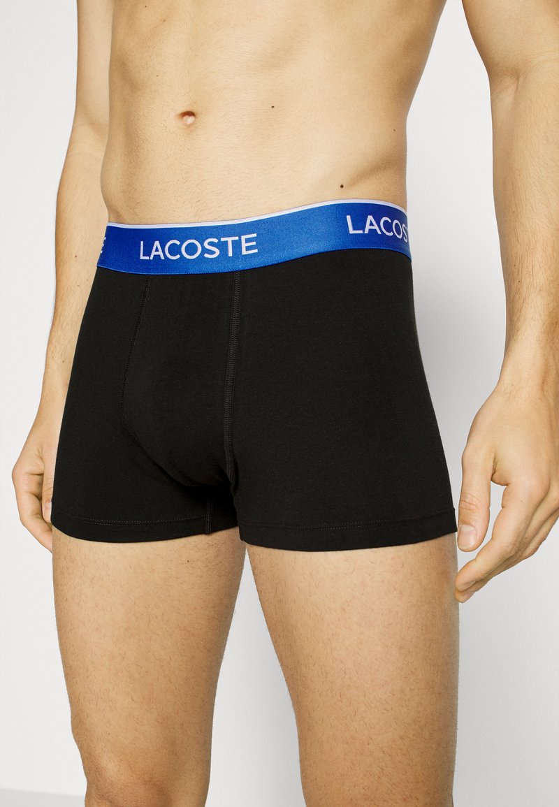 Boxer cortos negros con una cinturilla elástica azul que presenta el logo "LACOSTE" en blanco. El material parece suave, con un diseño ajustado.