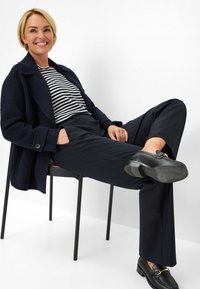 Marineblauwe wollen blazer, gestreepte zwart-witte top, donkere broek en zwarte loafers met gouden details. Zittend op een zwarte stoel tegen een witte achtergrond.