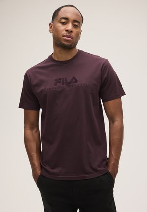 Giovane uomo in piedi con le mani nelle tasche, indossa una t-shirt FILA viola scuro contro uno sfondo semplice.