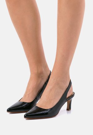 Piernas con zapatos de charol negro tipo slingback, con punta afilada y tacones de bloque de altura media sobre un fondo blanco.