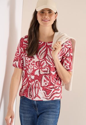 Femme souriante portant une casquette beige, une chemise à motifs floraux rouges et blancs, un jean bleu, tenant une veste beige clair sur l'épaule devant un fond uni.