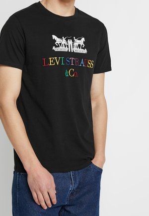 Mand iført sort Levi Strauss t-shirt med farverigt logo og blå jeans, stående med den ene hånd i lommen.