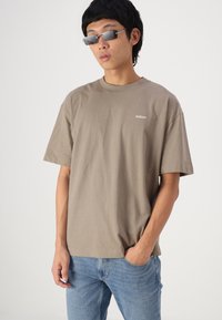 Νέος άνδρας που φοράει γκρι μπλούζα Hollister, ανοιχτό μπλε τζιν και μικρά ορθογώνια γυαλιά ηλίου, με το ένα χέρι στην τσέπη, σε ουδέτερο φόντο.