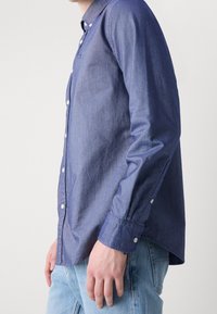 Camicia in denim blu scuro con bottoni bianchi, maniche lunghe e orlo curvo. Il tessuto appare liscio con una leggera lucentezza.