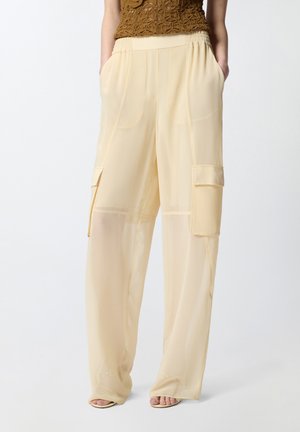 Donna che indossa pantaloni cargo beige trasparenti a gamba larga con tasche applicate, vita elastica e un top marrone a maglia senza maniche.