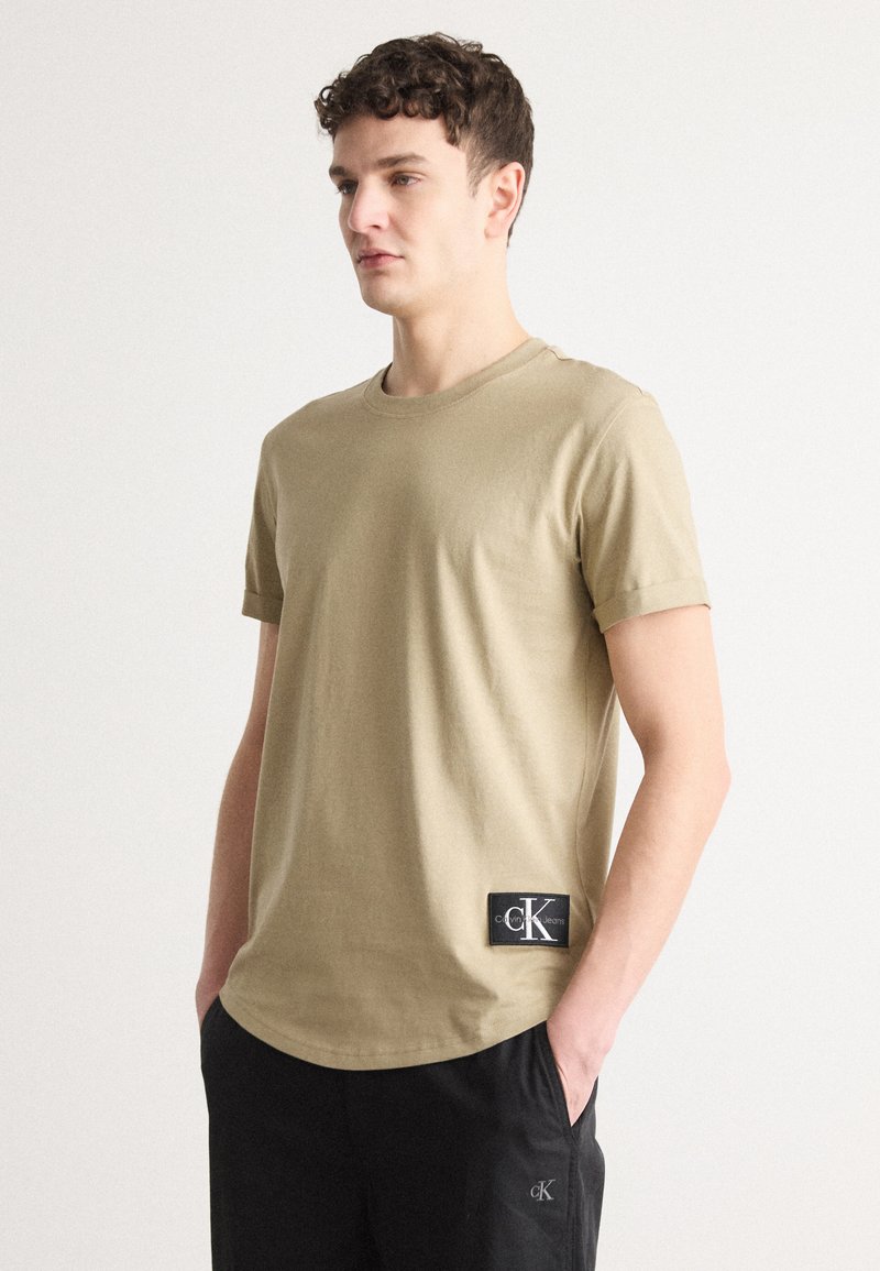 Tricou bej cu mânecă scurtă, dintr-un amestec moale de bumbac, având o etichetă cu logo-ul CK negru pe partea stângă jos, cu un guler clasic rotund.