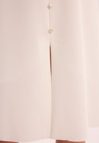 Kremowa sukienka z gładkiego, teksturowanego materiału. Ma rozcięcie i ozdobne guziki po lewej stronie. Prosty i elegancki design.