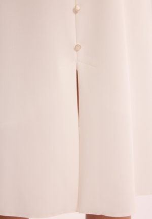 Kremowa sukienka z gładkiego, teksturowanego materiału. Ma rozcięcie i ozdobne guziki po lewej stronie. Prosty i elegancki design.