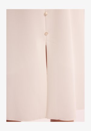 Kremowa sukienka z gładkiego, teksturowanego materiału. Ma rozcięcie i ozdobne guziki po lewej stronie. Prosty i elegancki design.