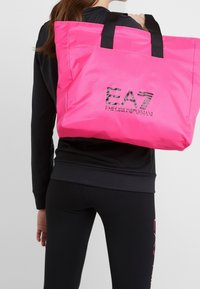 Růžová nylonová taška přes rameno s černým logem, vybavená dvěma textilními uchy a obdélníkovým tvarem. Hladká textura s minimalistickým designem.