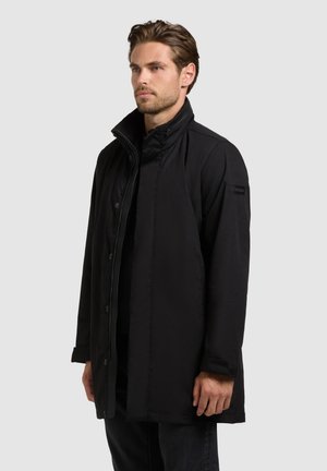 Man met kort bruin haar, draagt een zwarte jas met knopen en een hoge kraag, staand tegen een effen lichtgrijze achtergrond.