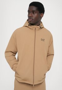 Veste beige zippée avec capuche, fabriquée en tissu lisse. Elle comprend un cordon de serrage réglable et un petit logo sur la poitrine.