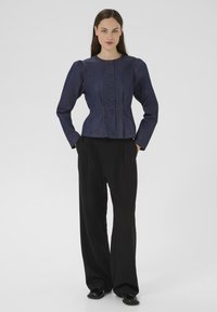 Giacca in denim scuro con silhouette aderente, scollo rotondo e maniche a sbuffo. Presenta chiusure con bottoni e dettagli di cuciture visibili. Abbinata a pantaloni neri ampi.