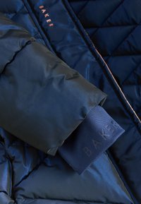 Manche de veste matelassée bleu foncé avec poignets noirs et nom de marque visible brodé sur le tissu.