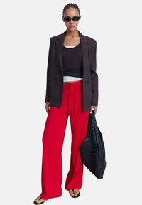 Rode brede pijpenbroek, zwart aansluitend topje, donkere oversized blazer en zwarte gevlochten draagtas. Accessoires omvatten zwarte zonnebril.