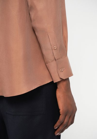 Braunes kurzärmliges Hemd mit glatter Textur, geknöpften Manschetten und abgerundetem Saum, kombiniert mit dunklen Hosen, die eine quadratische Tasche aufweisen.