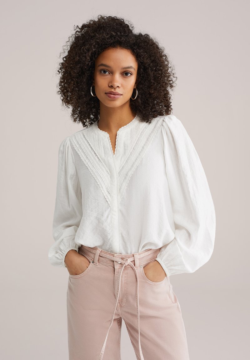 Blouse blanche texturée avec un col pointu, une patte de boutonnage et des plis verticaux ; manches bouffantes. Associée à un pantalon taille haute rose clair.